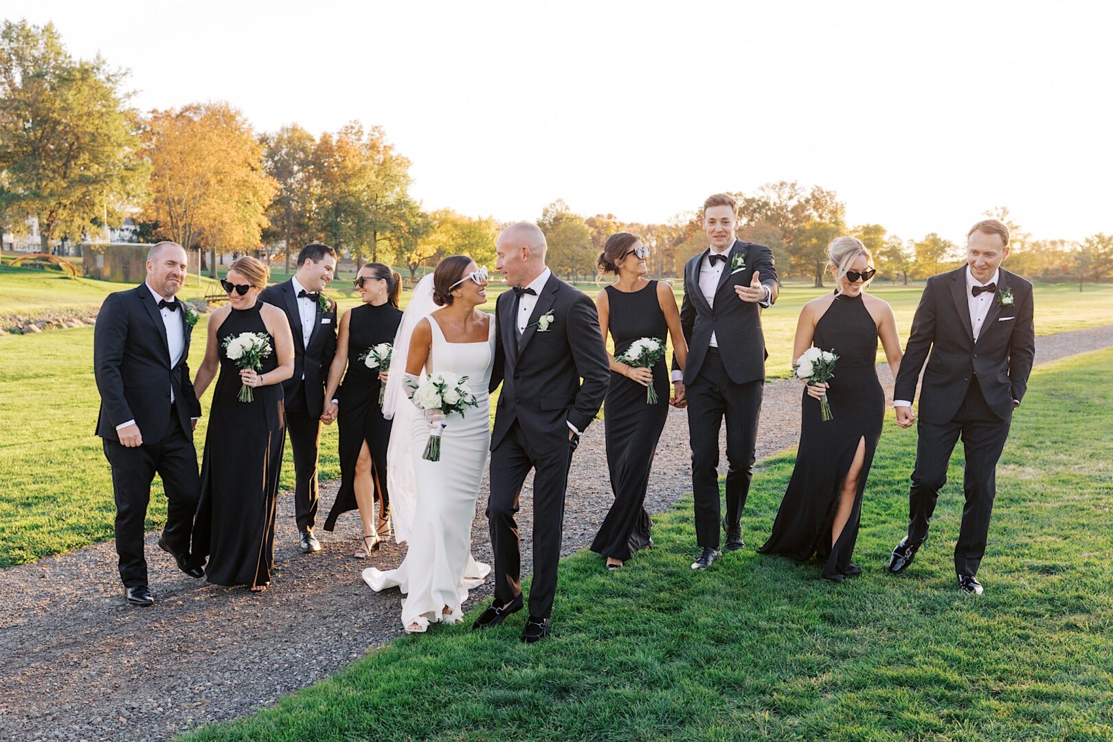 At The Tilling House wedding, a bride and groom stroll with their wedding party along a grassy path. The group, elegantly dressed in formal black and white attire, carries white bouquets. Majestic trees and a clear sky create the perfect backdrop for this special day.