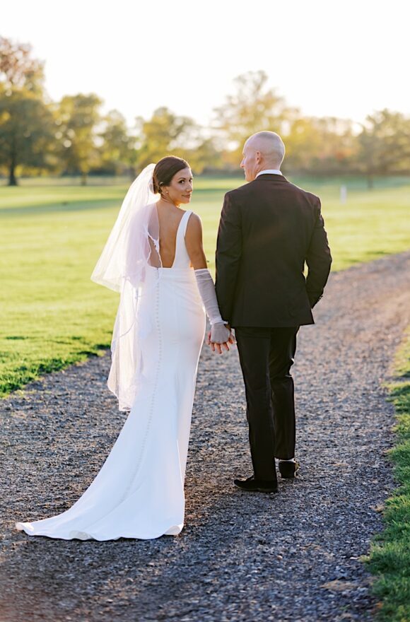 The bride and groom, having just celebrated their Tilling House wedding, walk hand in hand on a gravel path through a sunny park, with trees and grass creating the perfect romantic backdrop.