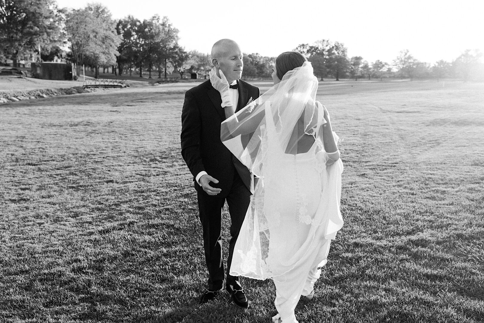 Bride and groom share a moment on a grassy field at The Tilling House wedding, with the sun setting in the background. The bride holds her veil as it flows behind her.