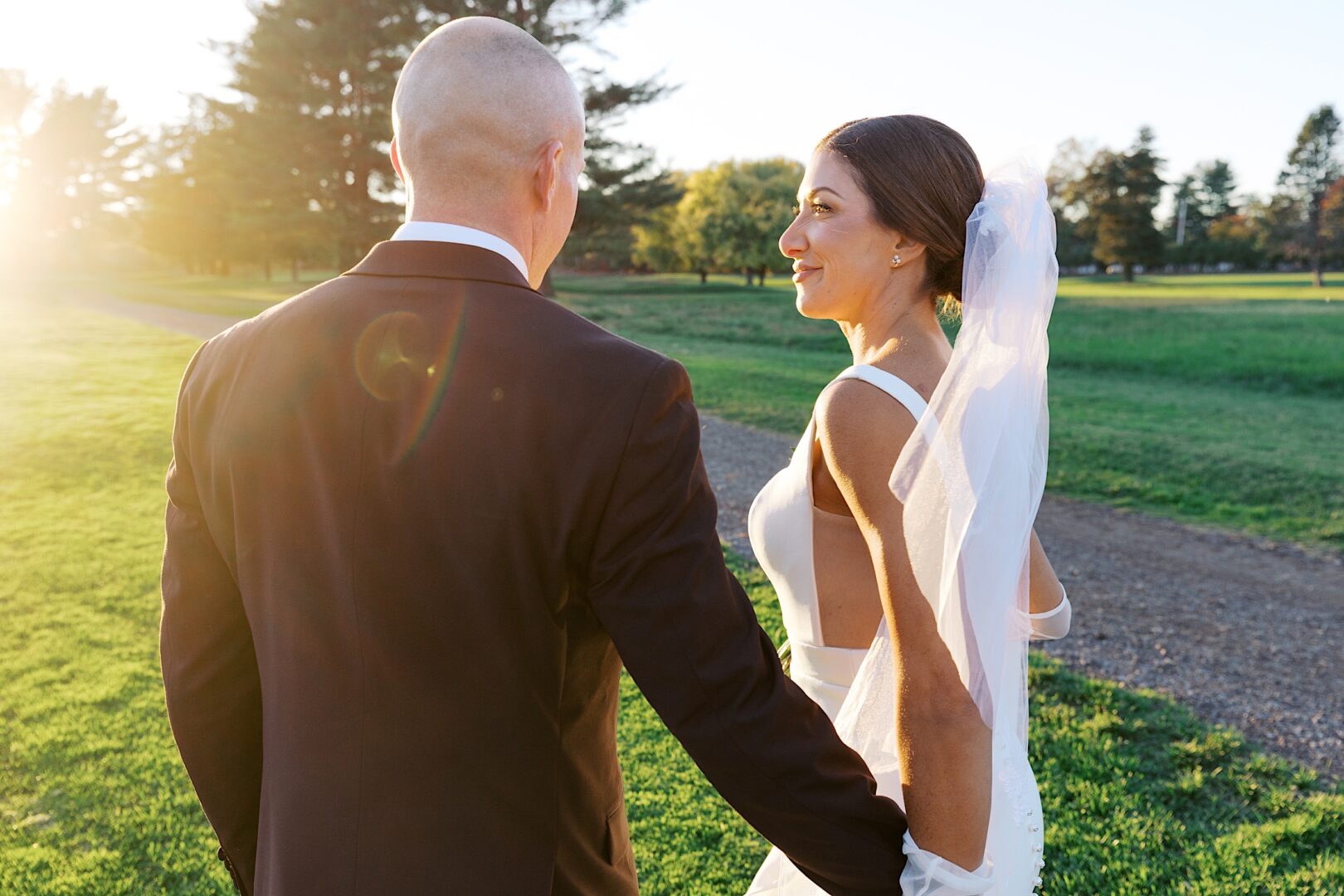 A couple in wedding attire walks hand in hand on a grassy path at sunset, with trees in the background, celebrating their Tilling House wedding. The bride gracefully wears a veil.