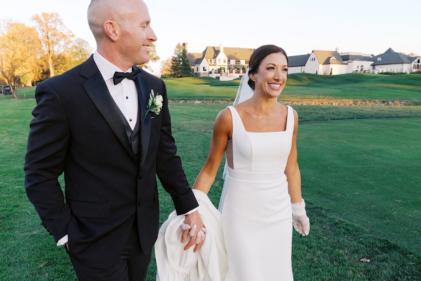 The bride and groom walk hand in hand on a grassy field, the elegance of The Tilling House wedding reflected in the picturesque buildings in the background.