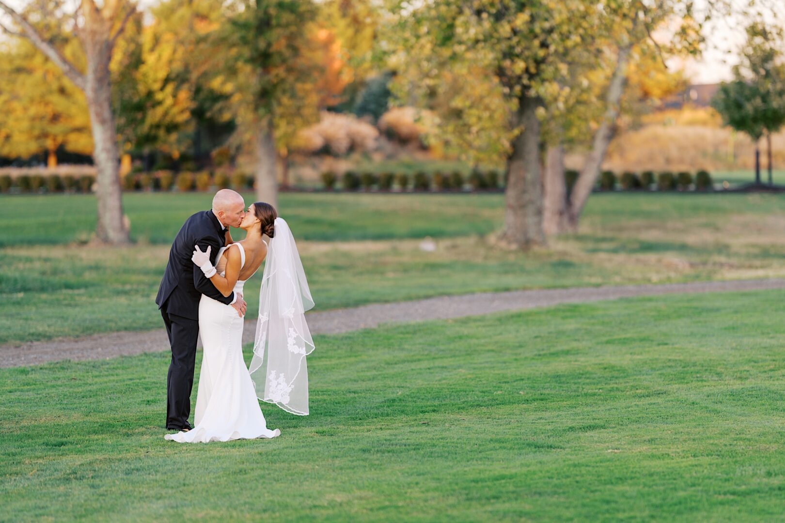 At The Tilling House wedding, a bride and groom share a tender kiss on a lush green lawn framed by majestic trees.