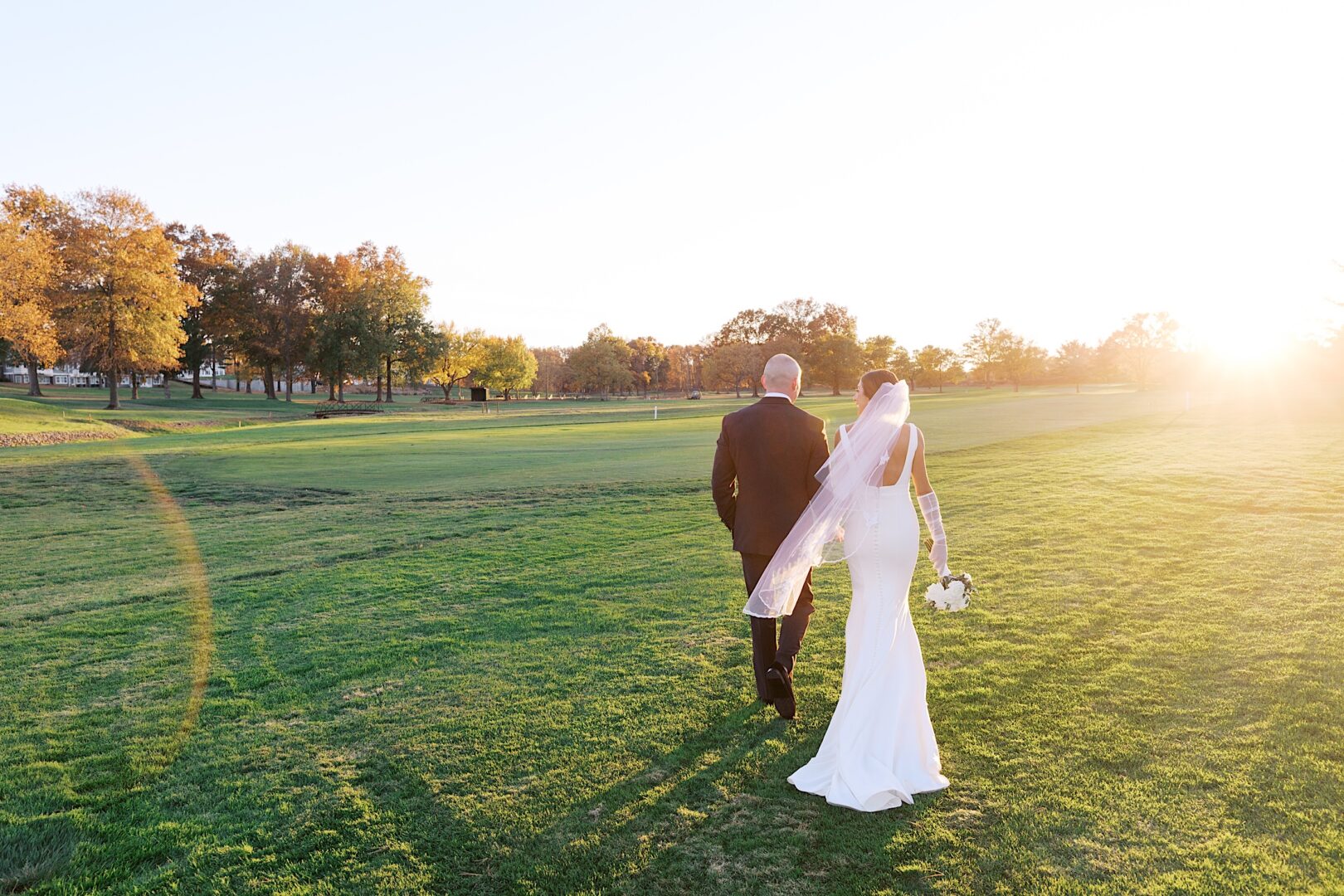 A bride and groom walk across a grassy field at The Tilling House wedding during sunset, with trees and a clear sky in the background. The bride holds a bouquet, and her veil flows elegantly behind her.