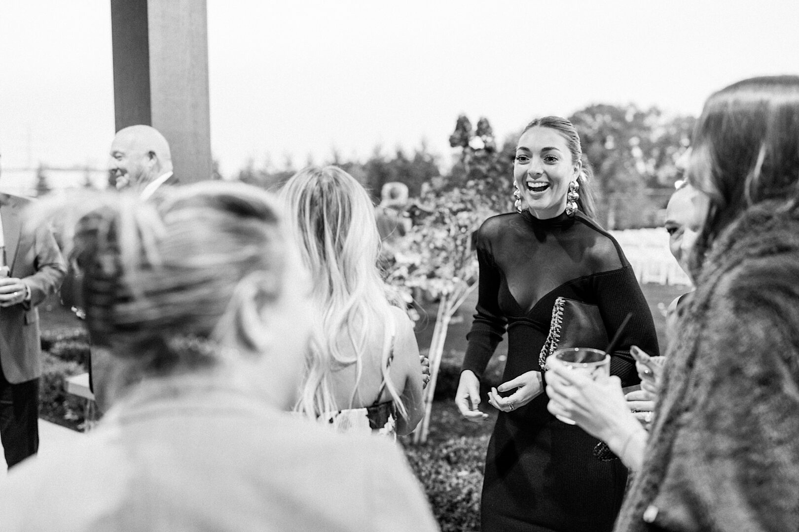 A group of people are socializing outdoors, a few holding drinks, at The Tilling House wedding. In the center, a woman in a black dress smiles radiantly. The entire scene is beautifully captured in black and white.