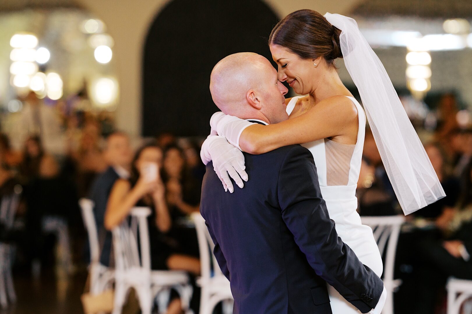 At The Tilling House wedding, a bride and groom share a joyful embrace during their dance. The bride dons a veil and gloves, while the groom looks sharp in his dark suit.