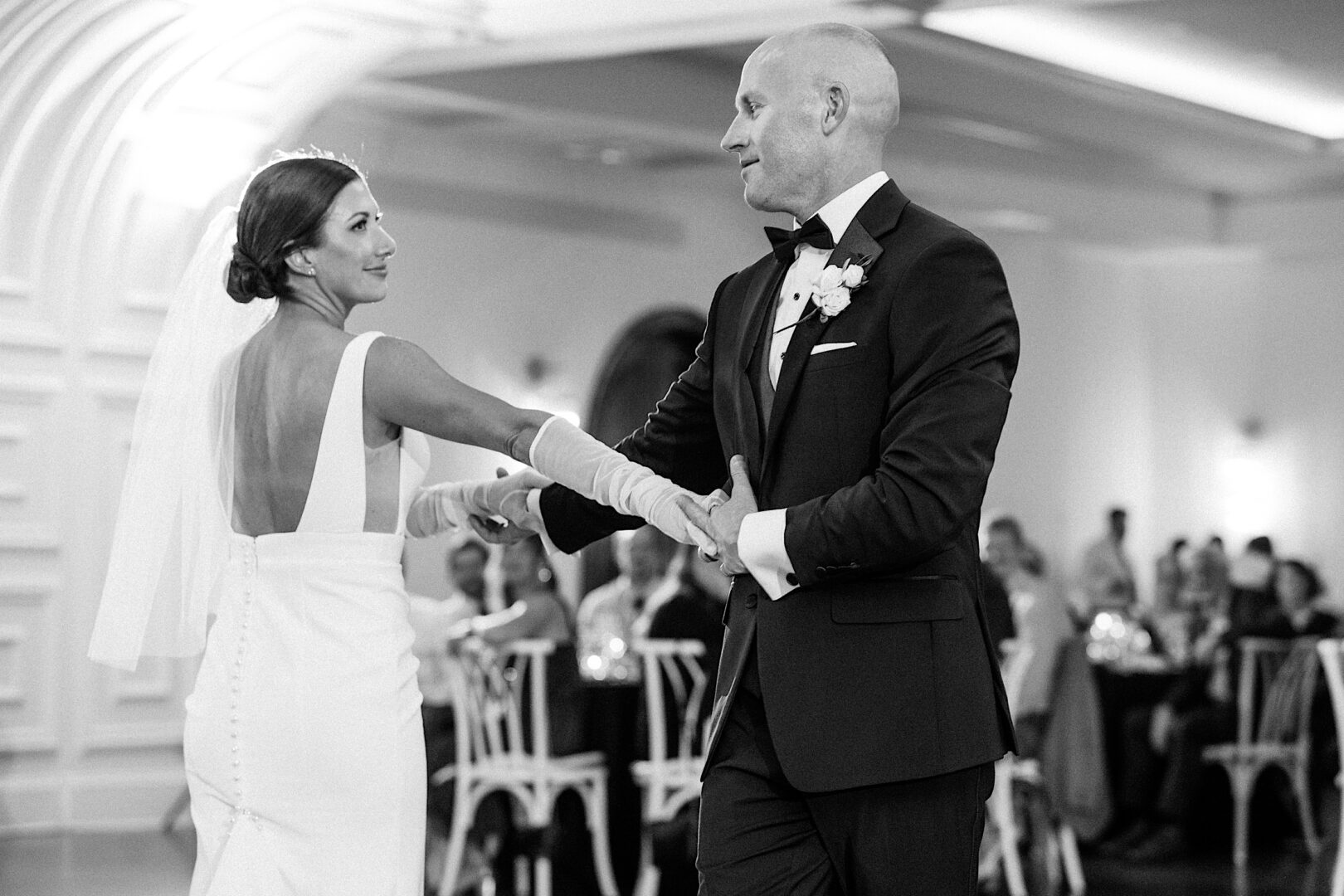 At The Tilling House wedding, a bride and groom share a dance in a spacious, well-lit room. The groom wears a tuxedo, and the bride dazzles in a white dress with a veil. Enthralled guests are seated in the background.