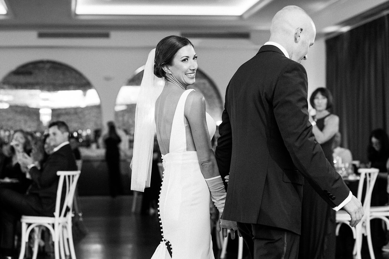 A bride and groom hold hands and walk together at The Tilling House wedding, with the bride smiling back at the camera. The background shows seated guests at tables, blurred.