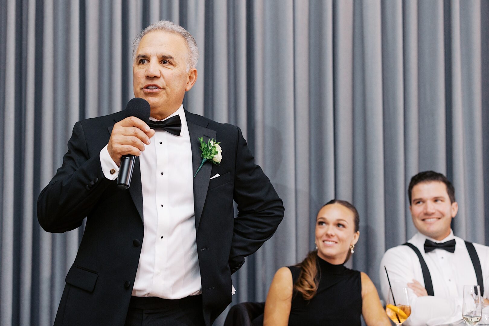 At The Tilling House wedding, a man in a tuxedo speaks into a microphone, standing in front of a gray curtain. A seated man and woman, both smiling, look towards him.
