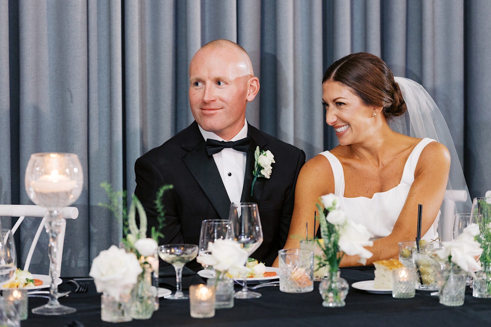 At The Tilling House wedding, a bride and groom sit at a table adorned with flowers and candles, smiling as they gaze to the right.