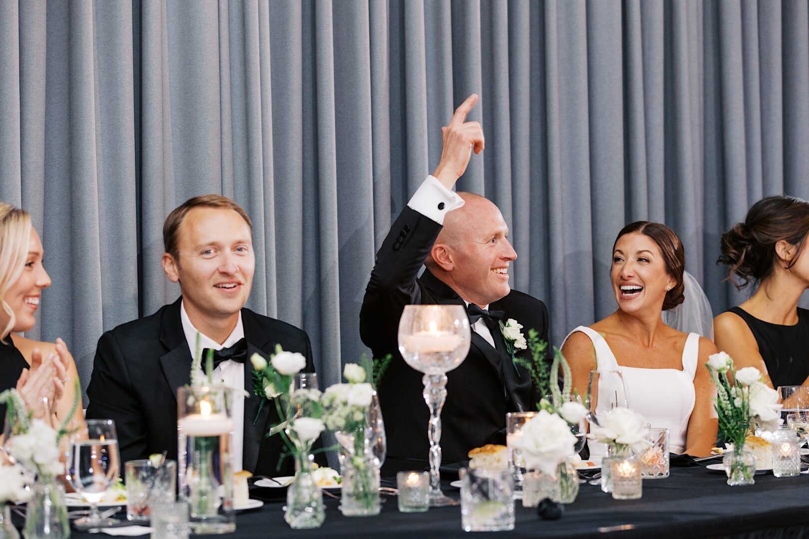 Guests in formal attire sit at a table adorned with flowers and candles, celebrating The Tilling House wedding. A bald man raises his hand amid smiling faces, all set against a backdrop of gray curtains.