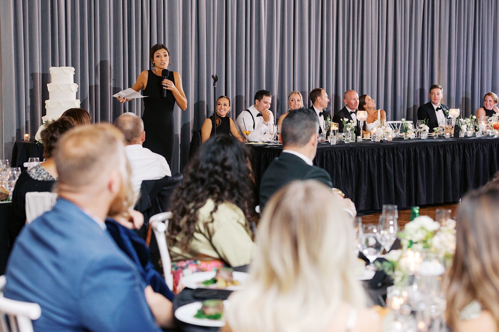 A person is speaking into a microphone at the Tilling House wedding, where guests are seated at long tables with a four-tiered cake in the background.