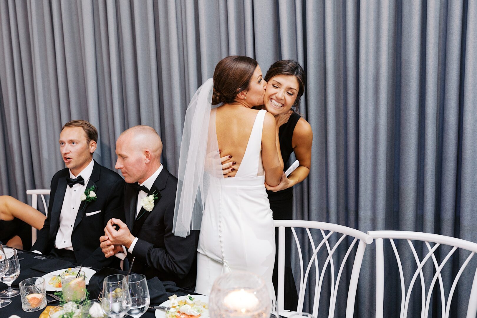 At The Tilling House wedding, a bride in a white dress hugs a woman in black, while two men in tuxedos sit at a table with empty plates and glasses.