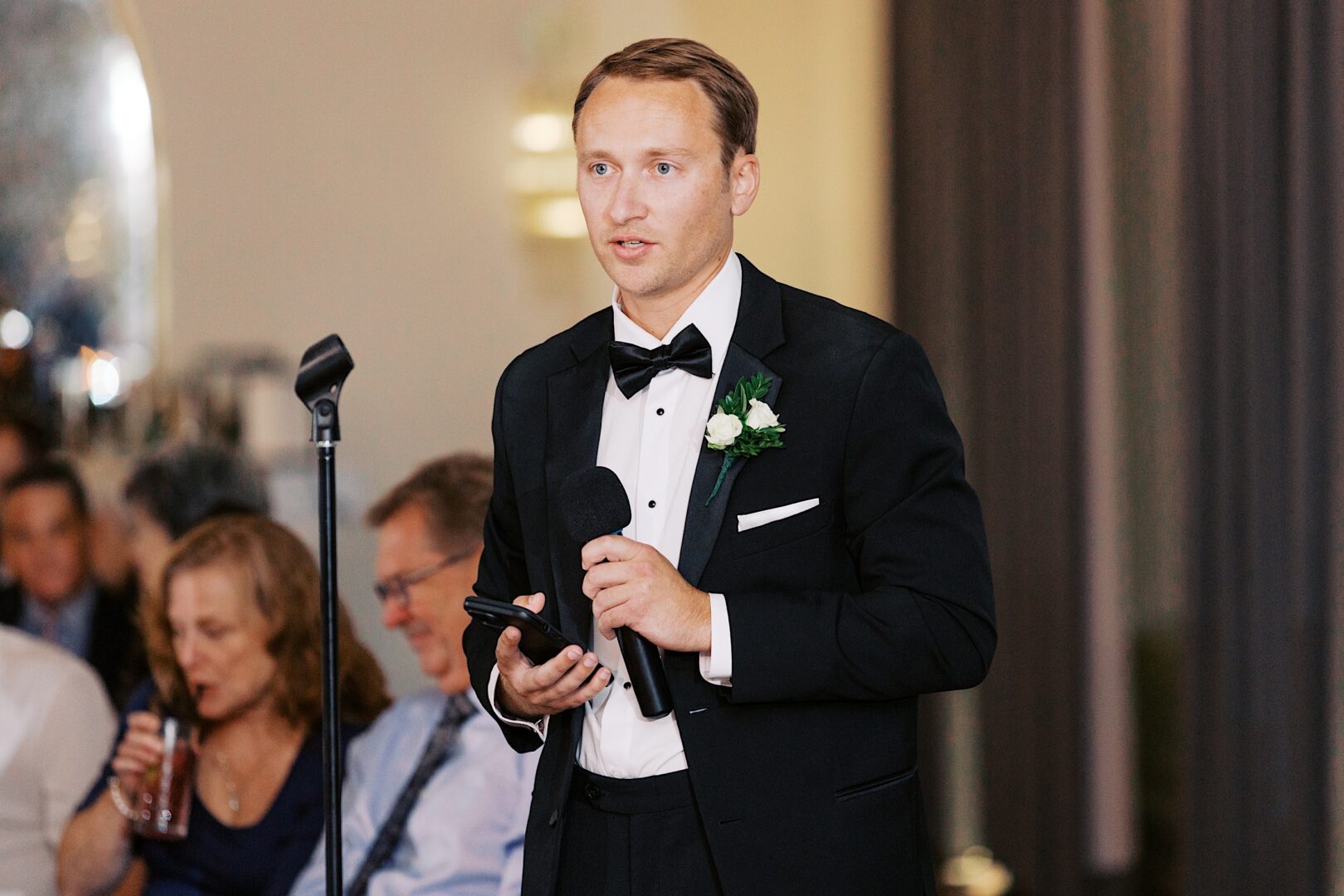 A man in a tuxedo speaks into a microphone at The Tilling House wedding, elegantly holding a phone in his hand during the indoor event.