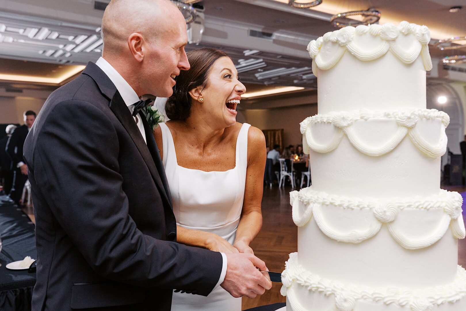 At The Tilling House wedding, a couple in formal attire shares laughter while cutting a tall, white-tiered wedding cake in the elegantly adorned banquet hall.