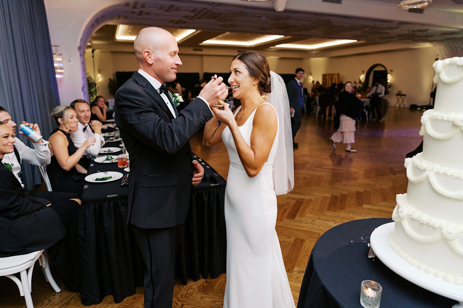 A couple in wedding attire at The Tilling House wedding lovingly feeds each other cake in a banquet hall, surrounded by seated guests. A multi-tiered cake takes center stage in the foreground.