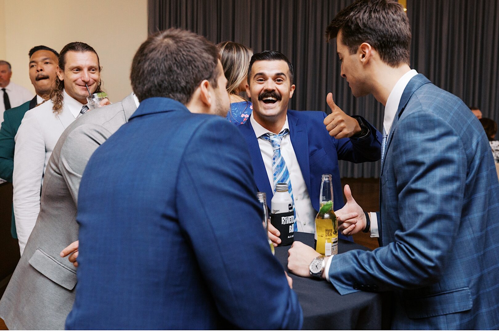 At The Tilling House wedding, a group of people in suits gathers around a cocktail table, socializing and holding drinks. One man is smiling and giving a thumbs-up, infusing the elegant event with a touch of casual charm.