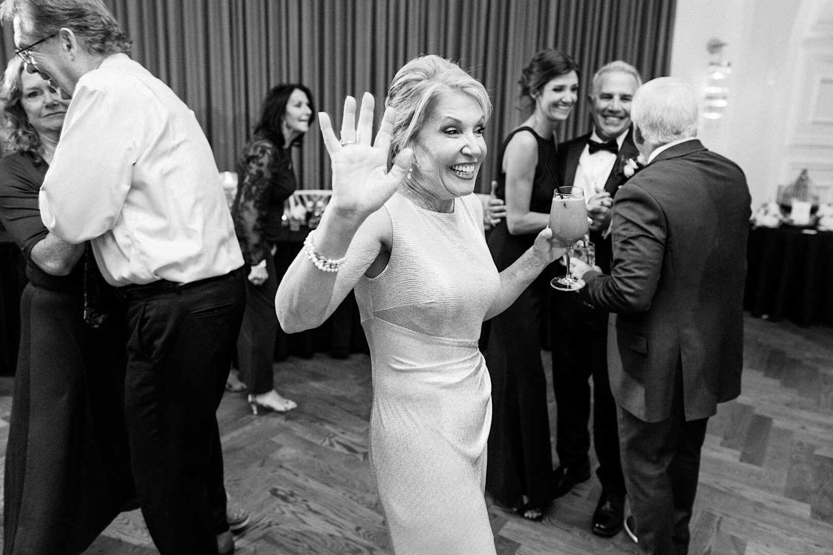 A woman in formal attire waves at the camera, capturing a joyful moment at The Tilling House wedding, while guests socialize in the background at an indoor event.