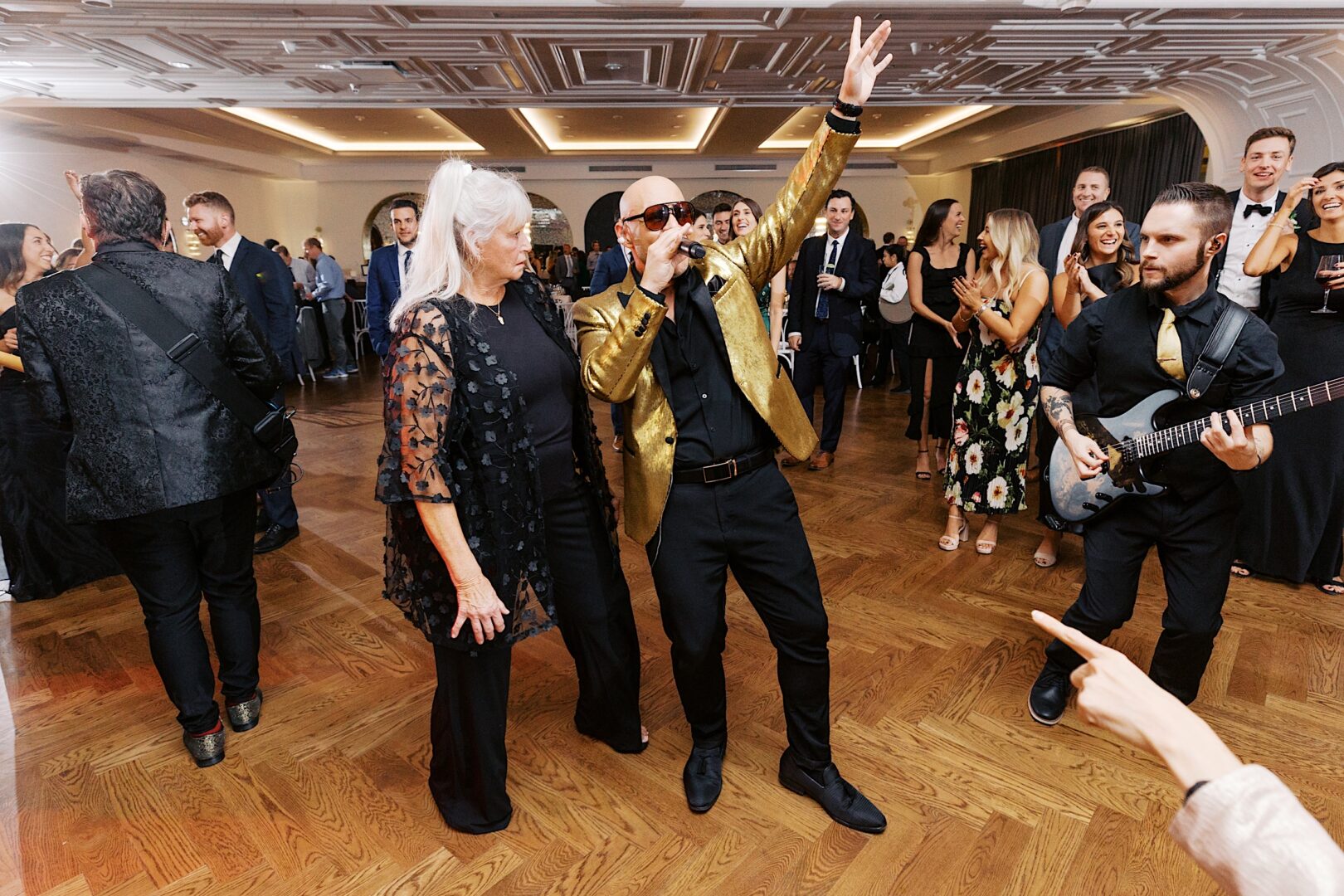 At The Tilling House wedding, a singer in a gold jacket performs energetically on the dance floor, surrounded by guests, including a woman in black and a guitarist.