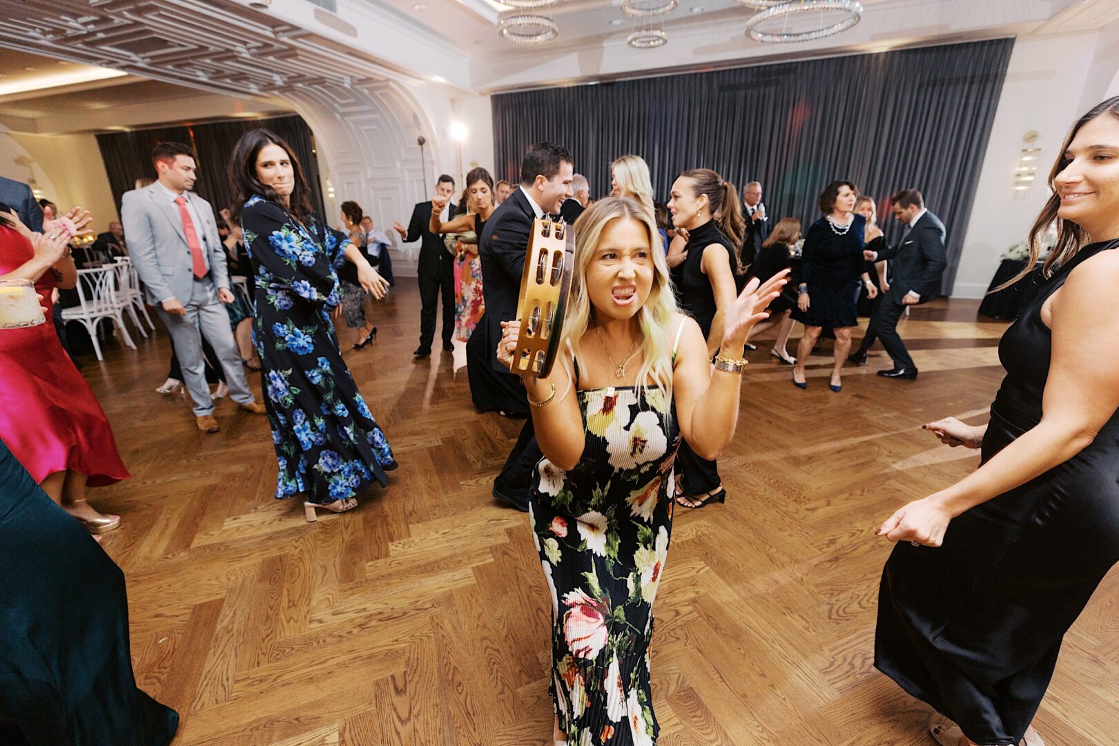 A lively group of people in formal attire dance on the wooden floor during The Tilling House wedding. In the foreground, a woman in a floral dress joyfully plays a tambourine.