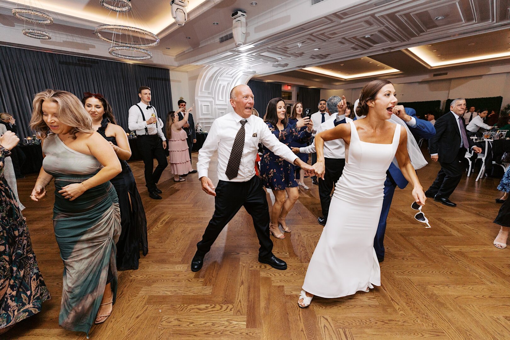 People dancing energetically on a wooden floor at The Tilling House wedding, with chandeliers and tables in the background.