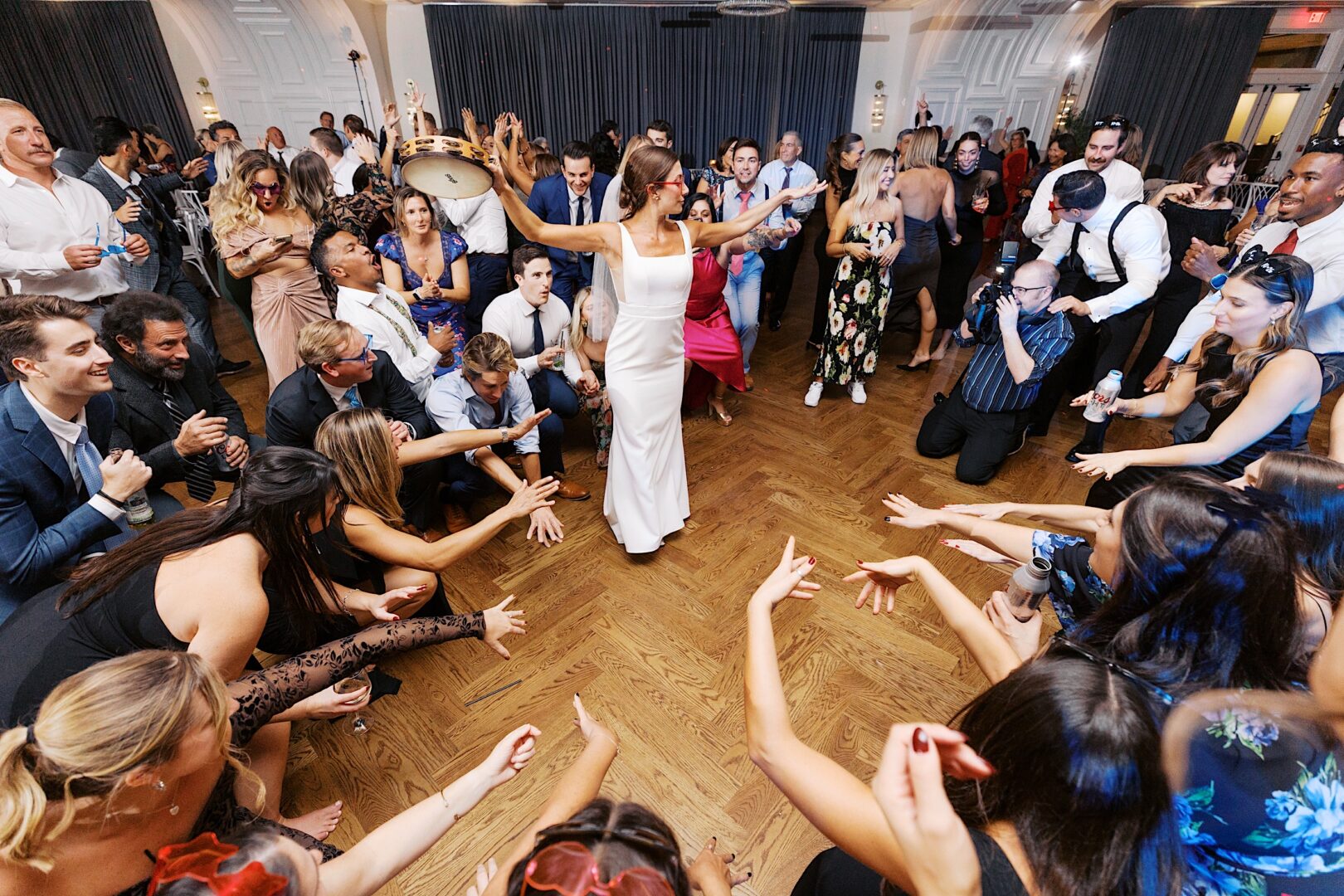 At The Tilling House wedding, a woman in a white dress dances gracefully at the center of a lively crowd, with guests clapping and reaching towards her on the wooden floor.