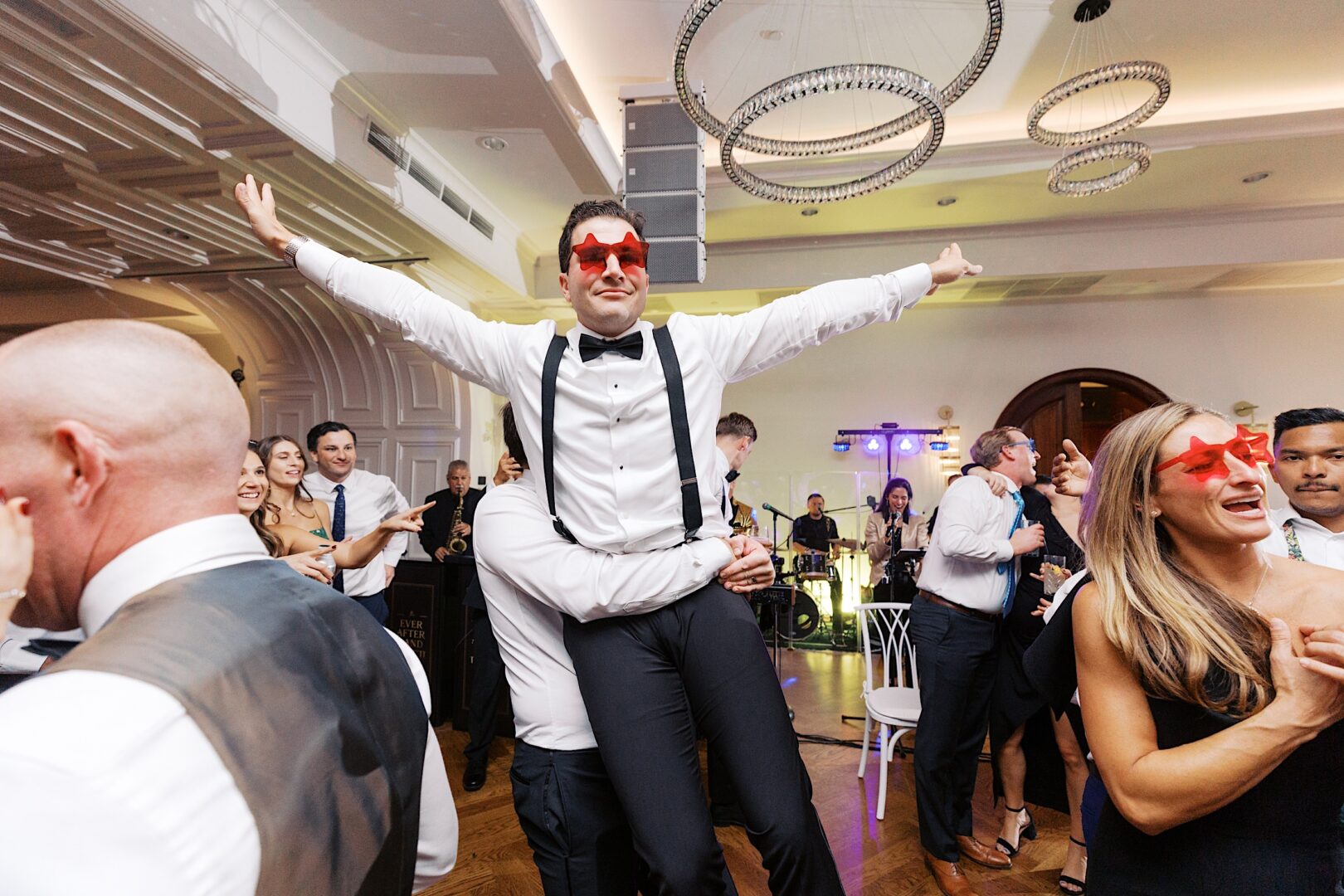 At The Tilling House wedding, a man in formal attire wearing red star-shaped glasses is joyfully lifted by another person amidst a lively indoor celebration where guests are dancing and the band is playing energetically.