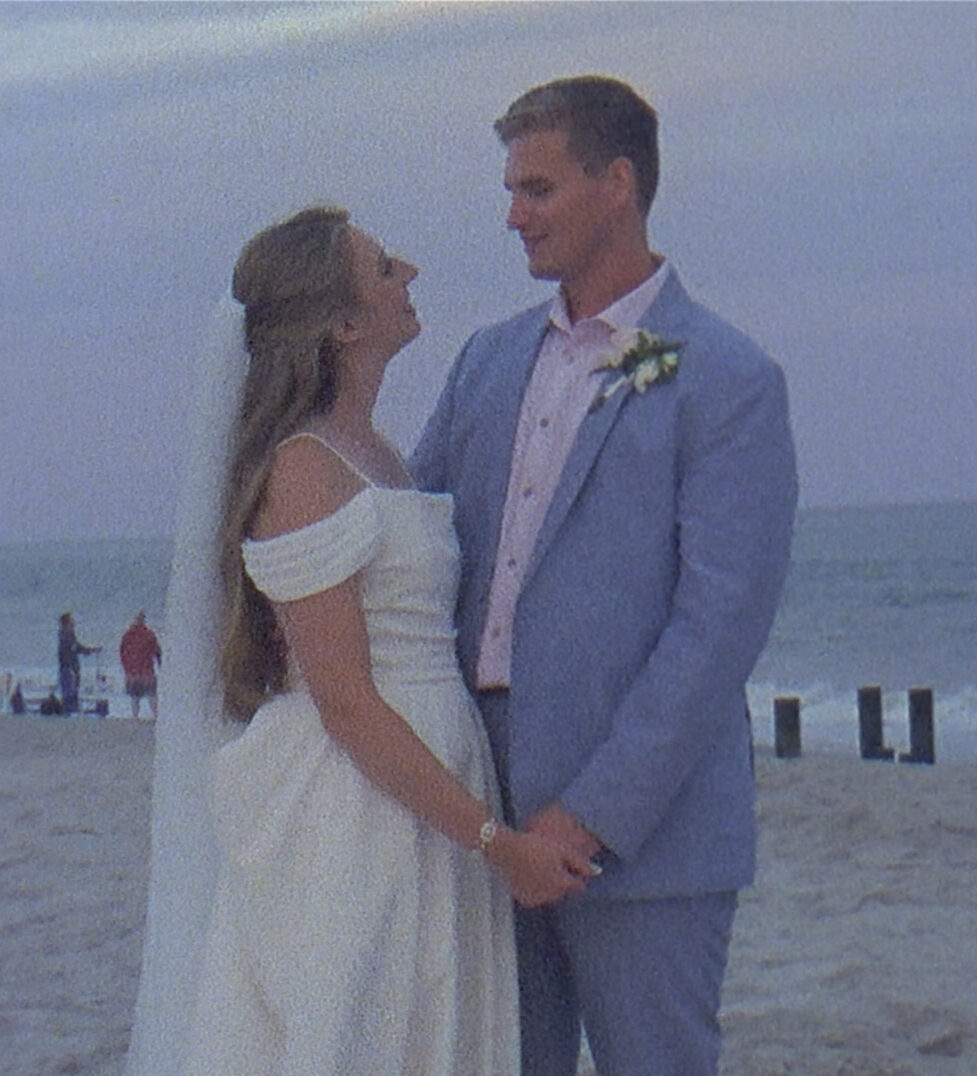 A couple in wedding attire stands on the beach, holding hands and gazing at each other, with the ocean stretching behind them during their enchanting Cape May wedding.