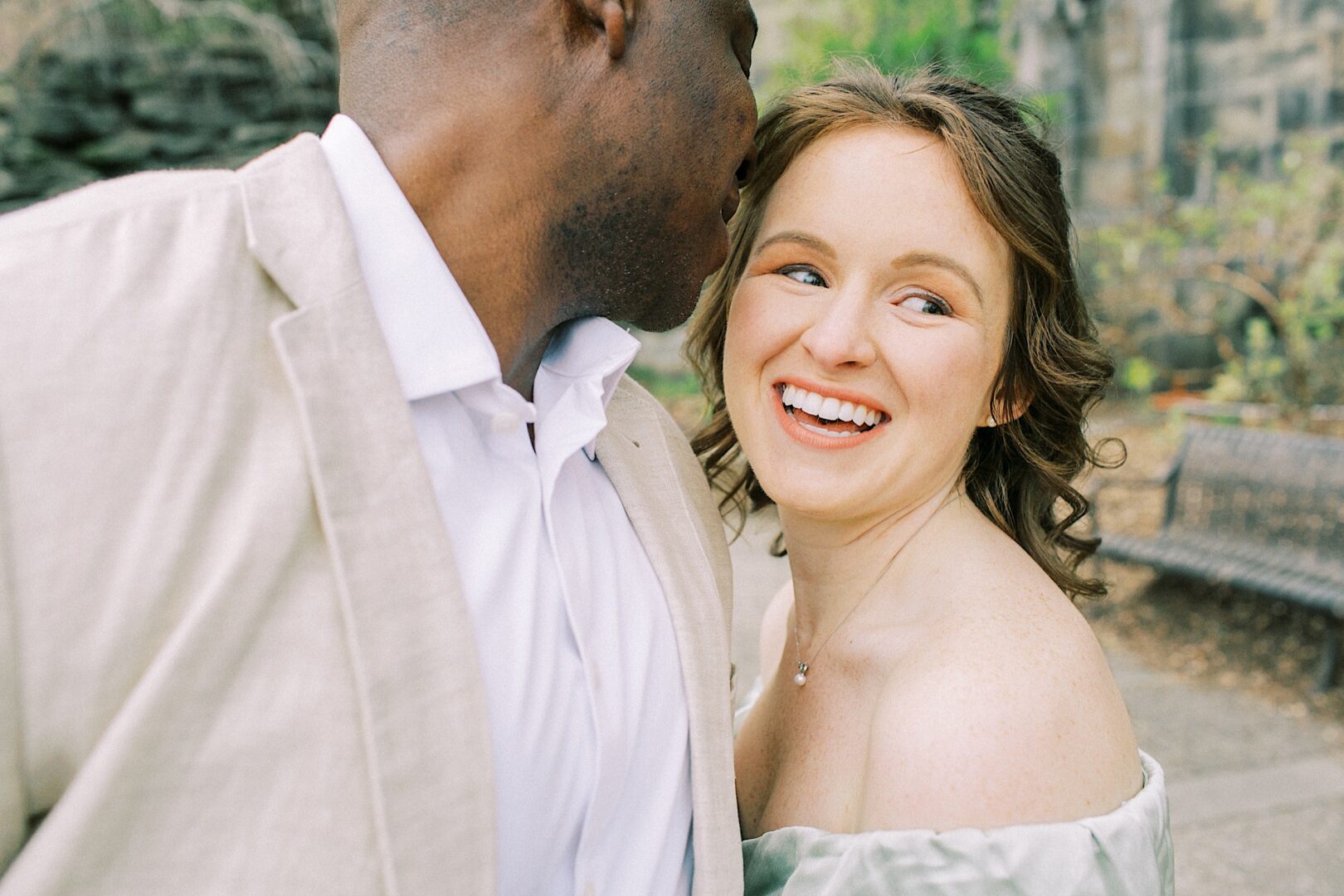 A woman smiling while a man whispers to her outside. The man is wearing a light suit, and the woman is in an off-shoulder dress. They are in a natural setting.