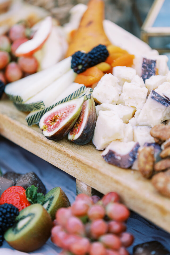 A wooden platter at a South Jersey wedding showcases assorted cheeses, figs, pear slices, blackberries, apricots, walnuts, grapes, strawberries, and kiwi.