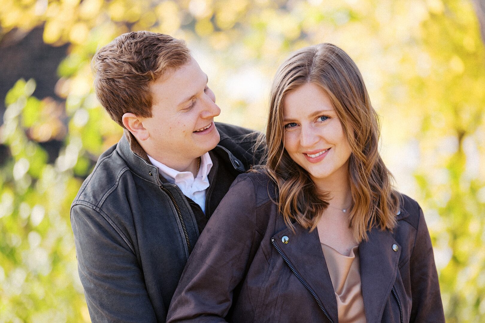 A couple stands together outdoors, both wearing jackets. The man is smiling at the woman, who is looking at the camera with a slight smile. The background features blurred greenery.