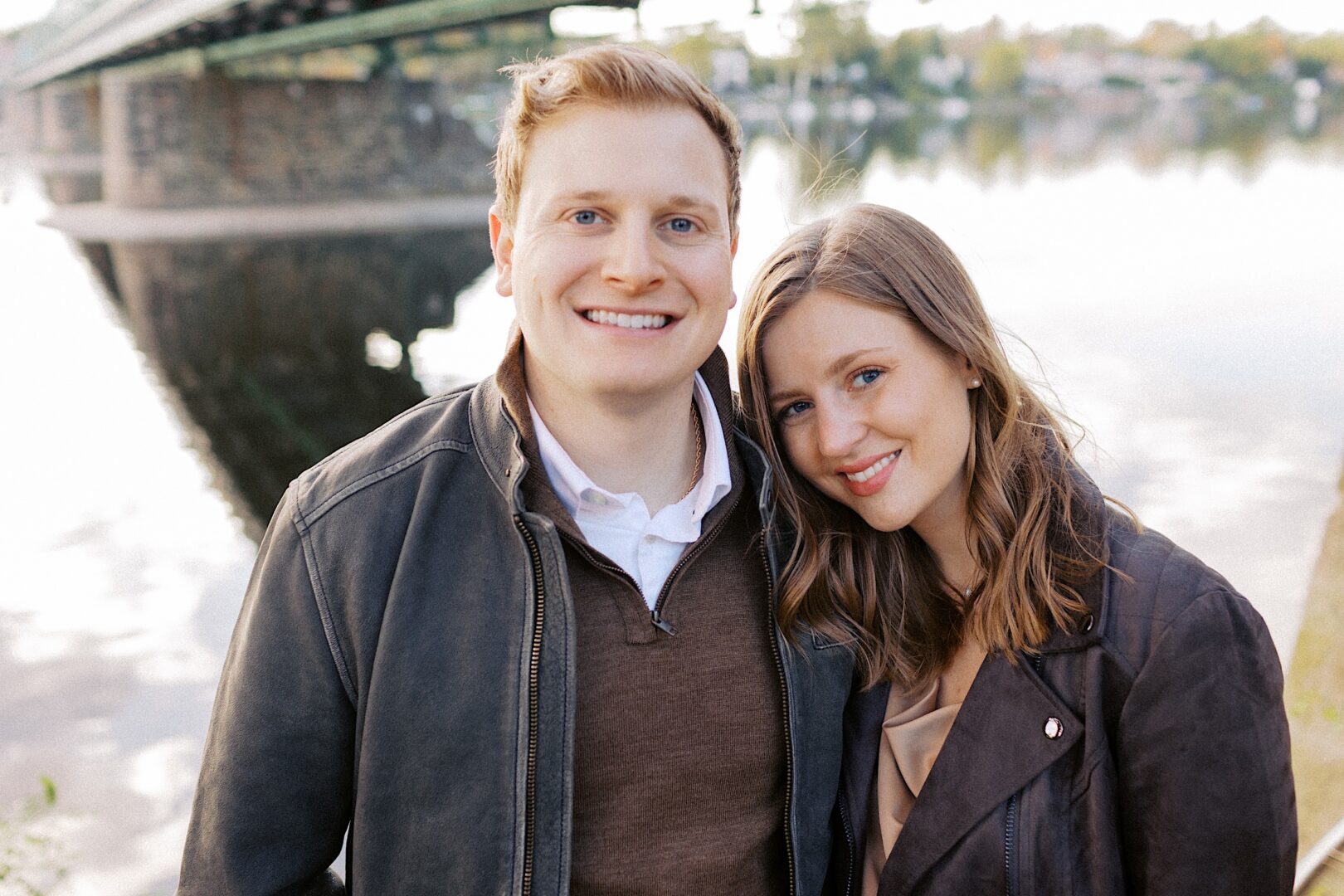A smiling man and woman stand closely together outdoors by a river, with a bridge in the background.