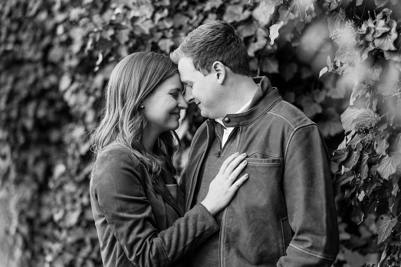 A couple stands closely, smiling and touching foreheads, in front of a leafy wall. The woman has her hand on the man's chest. Black and white photo.