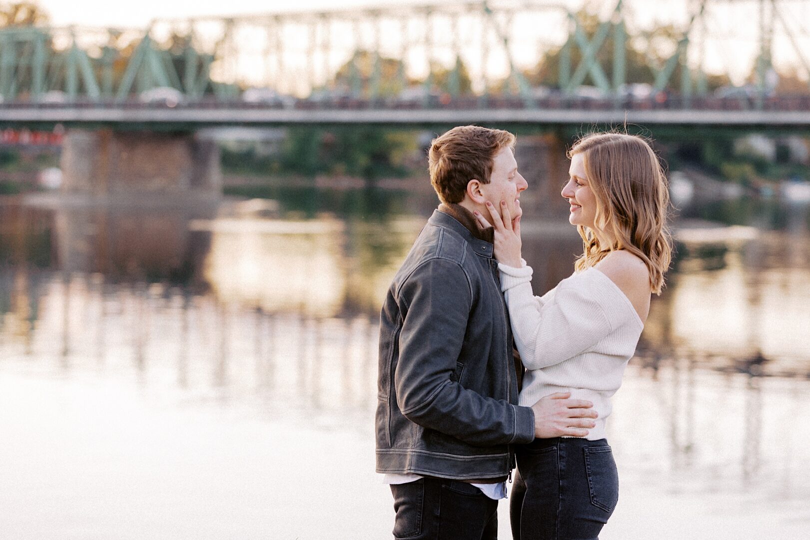 A couple stands close near a river, facing each other with a bridge in the background.