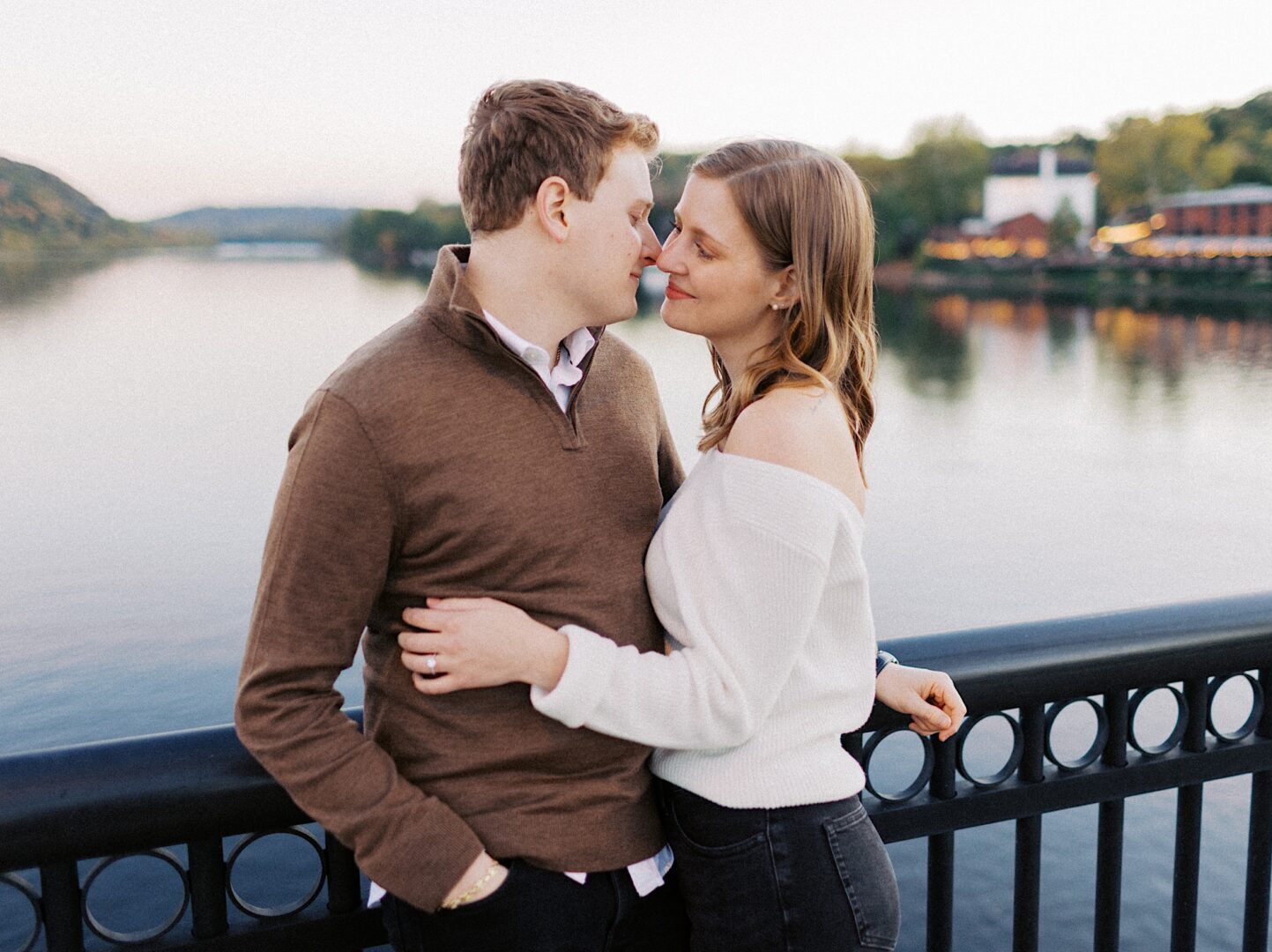 A couple stands on a bridge over a calm river, embracing and smiling at each other, with trees and buildings visible in the background.