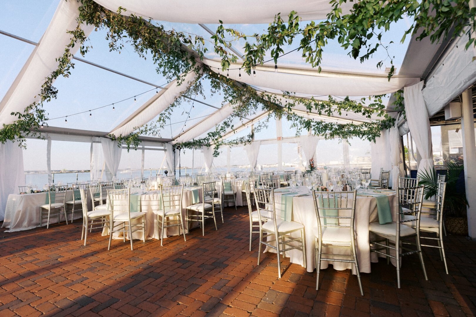 Outdoor wedding reception setup with round tables and white chairs under a transparent tent. Green foliage decorates the canopy, creating a picturesque scene perfect for South Jersey wedding photos. Brick flooring is visible, adding rustic charm to the elegant setting.