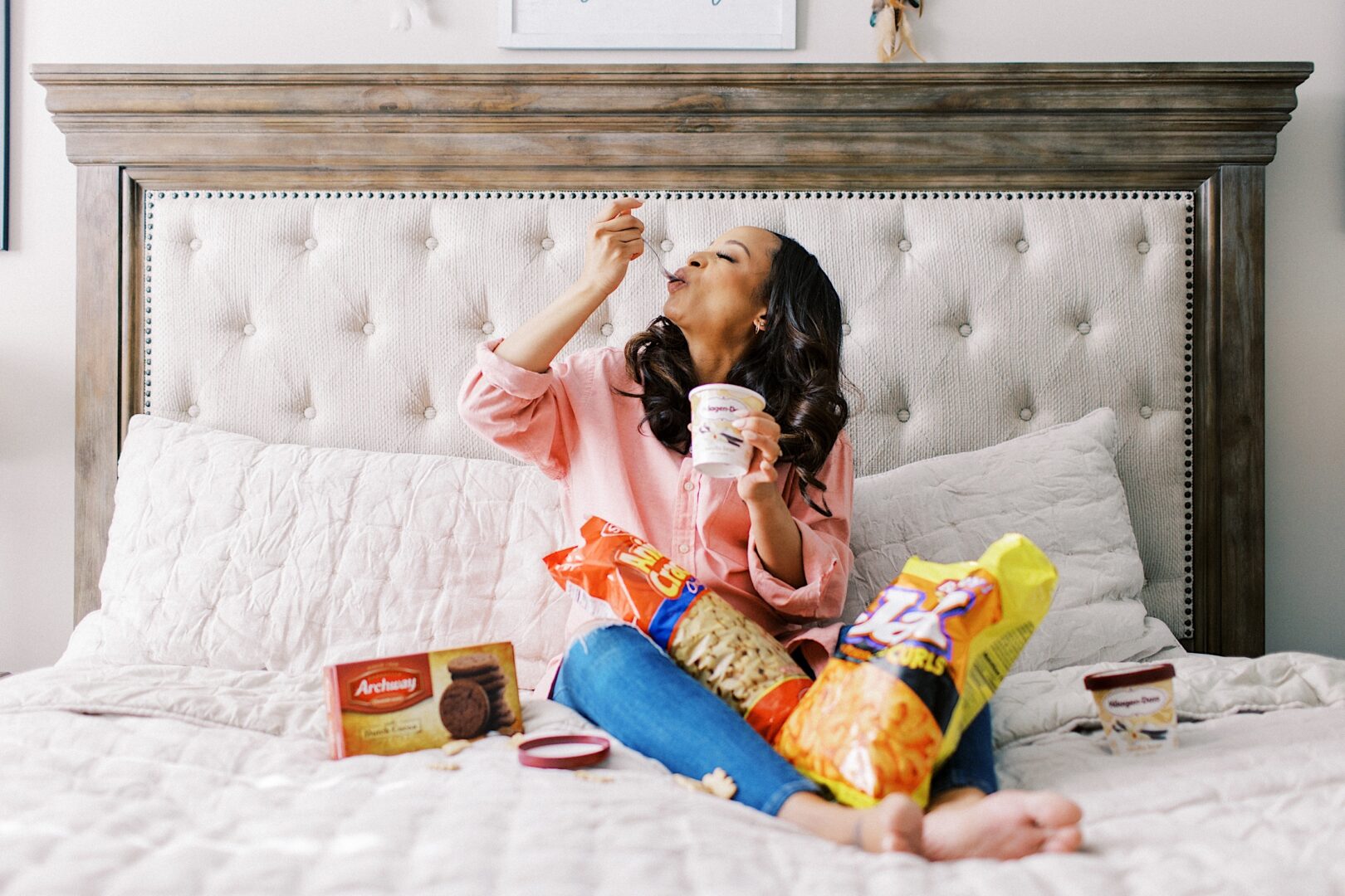 A pregnant woman in a pink shirt eating ice cream on a bed, surrounded by snacks, including chips and cookies.