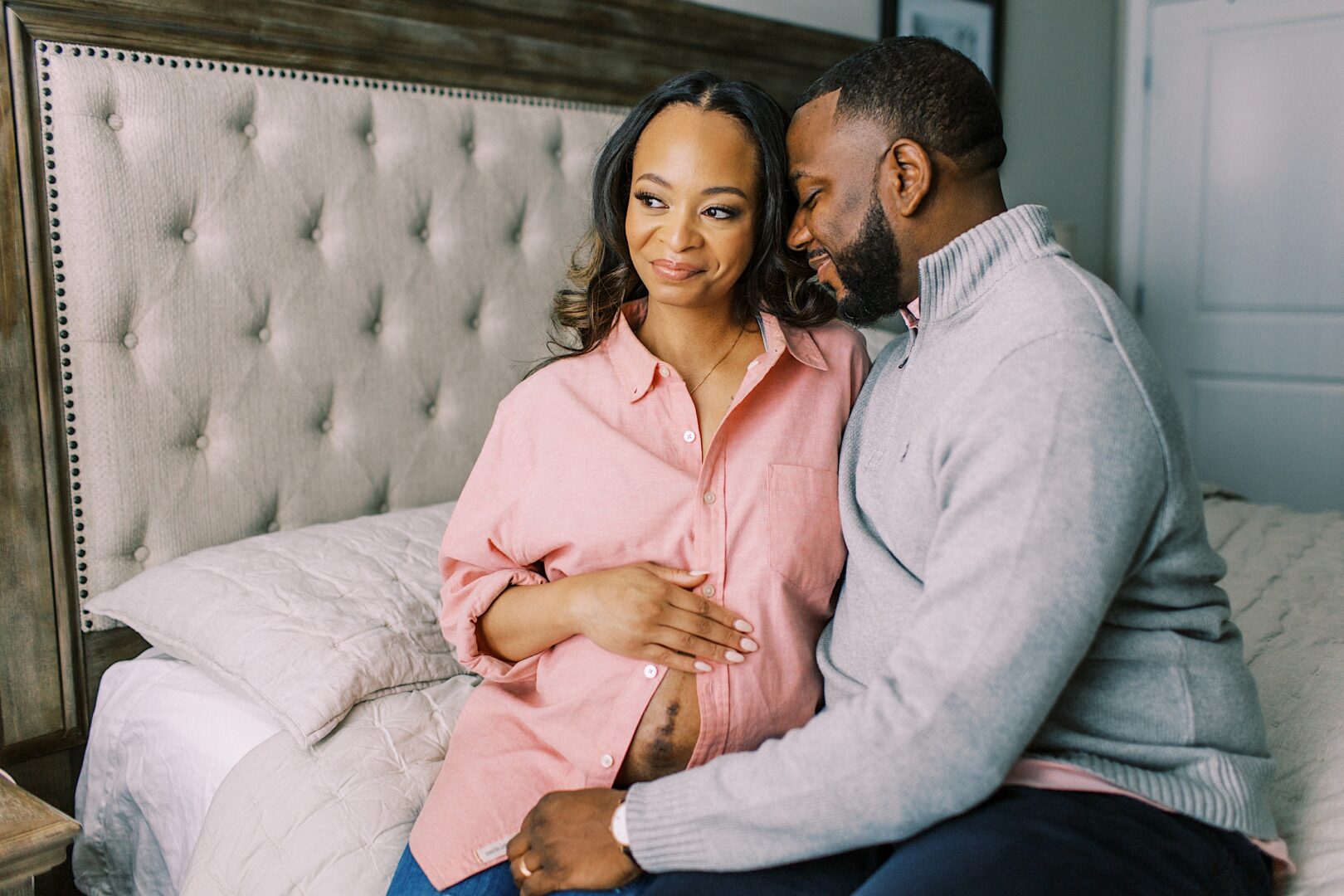 A couple sits on a bed; the woman in a pink shirt holds her pregnant belly, while the man in a gray sweater sits close to her, both displaying happy expressions.