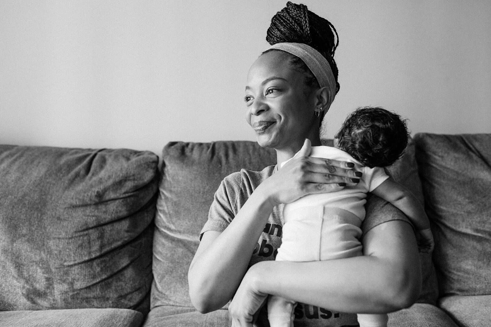 A woman sitting on a couch holds a baby over her shoulder. She looks to the side and smiles. The photo is in black and white.