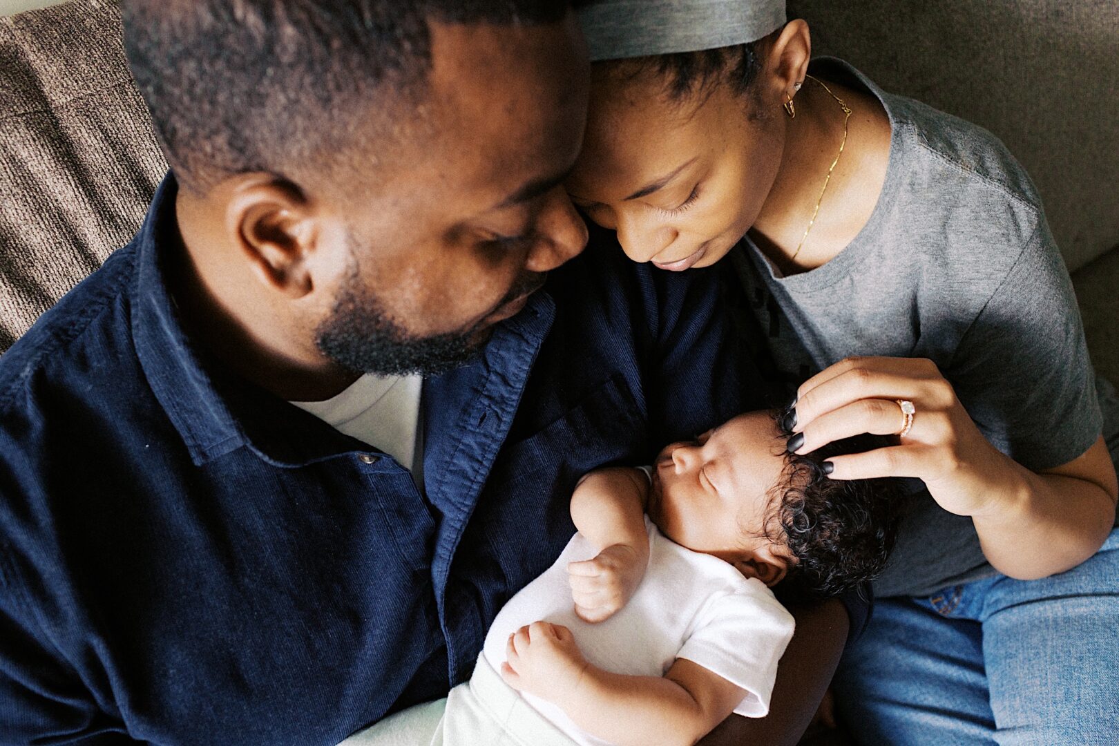A man and woman sit closely on a couch, looking at a sleeping baby in the man's arms. The woman gently touches the baby's hair.
