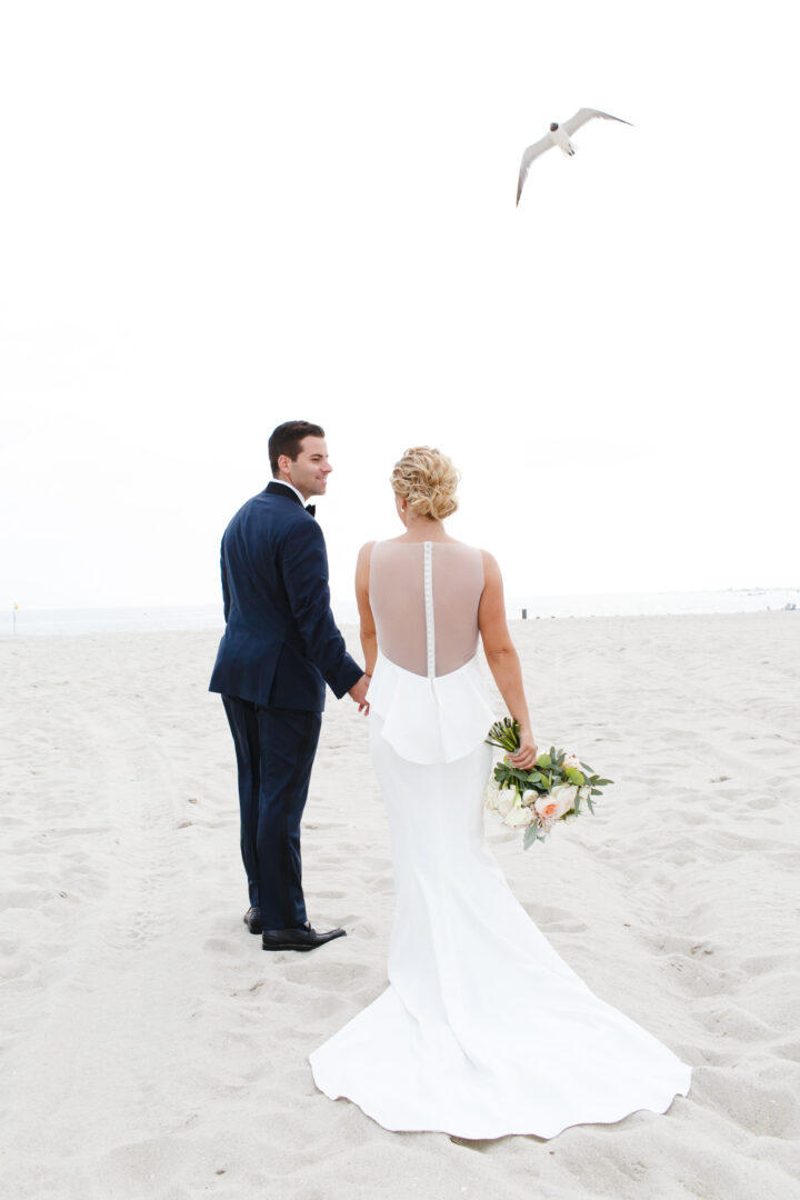 A couple in wedding attire holds hands on a South Jersey beach as a seagull flies overhead. The bride carries a bouquet, capturing the essence of timeless wedding photos.