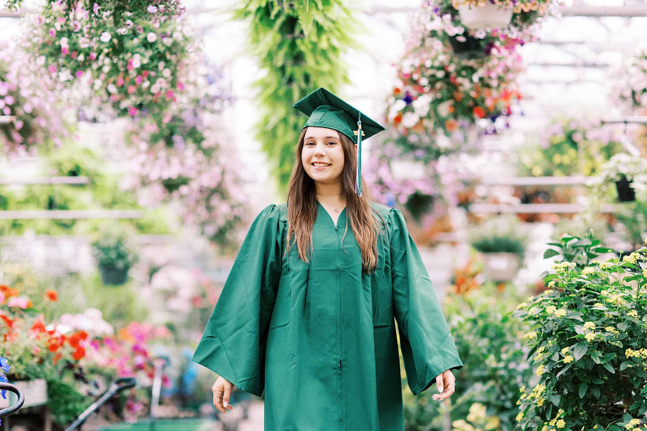 A person in a green graduation gown and cap stands smiling in a garden center surrounded by colorful flowering plants during their Senior Portrait Session in Montgomery County.