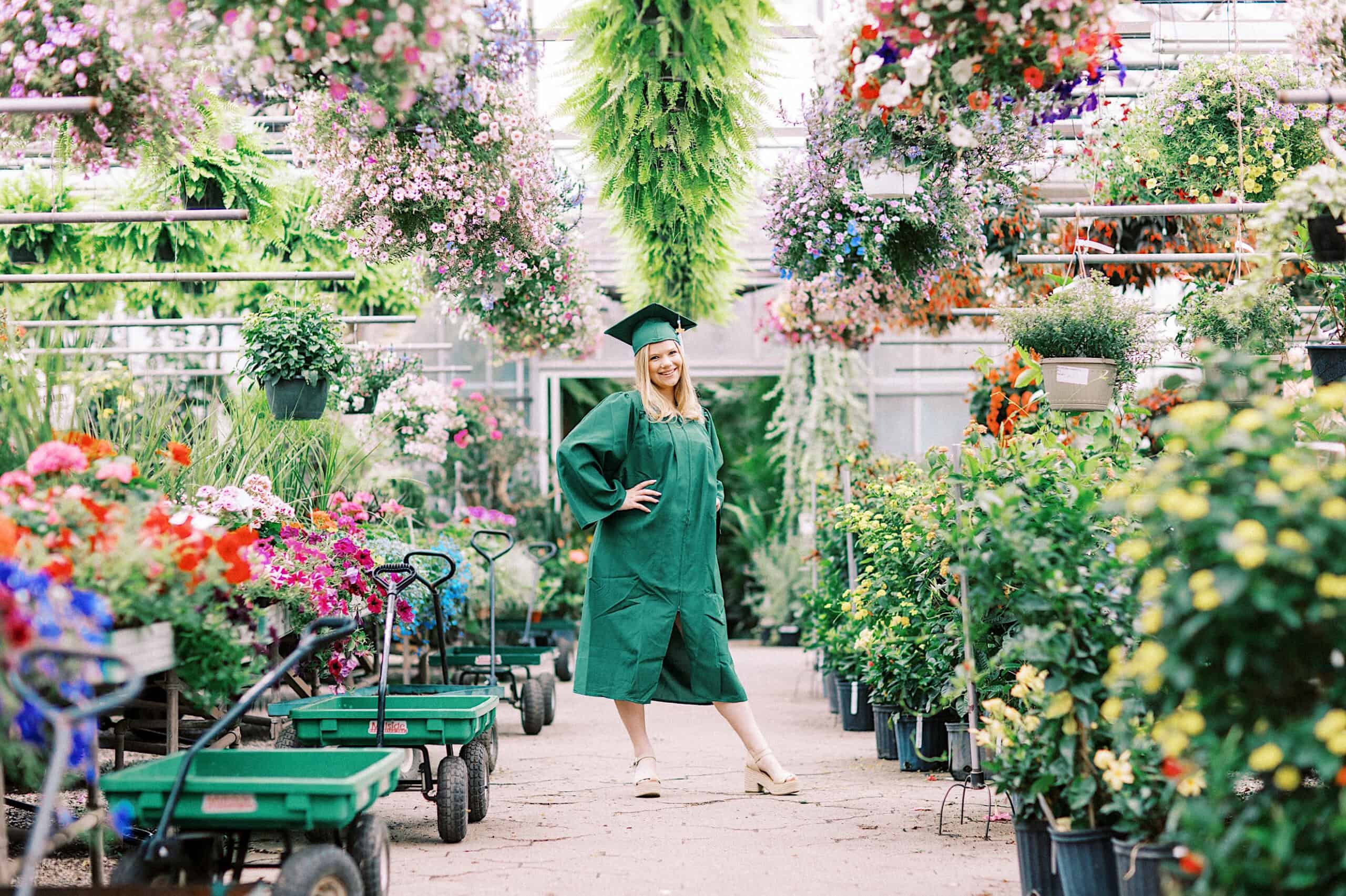 A person in a green graduation cap and gown stands smiling in a greenhouse surrounded by colorful flowers and plants during their Senior Portrait Session in Montgomery County.