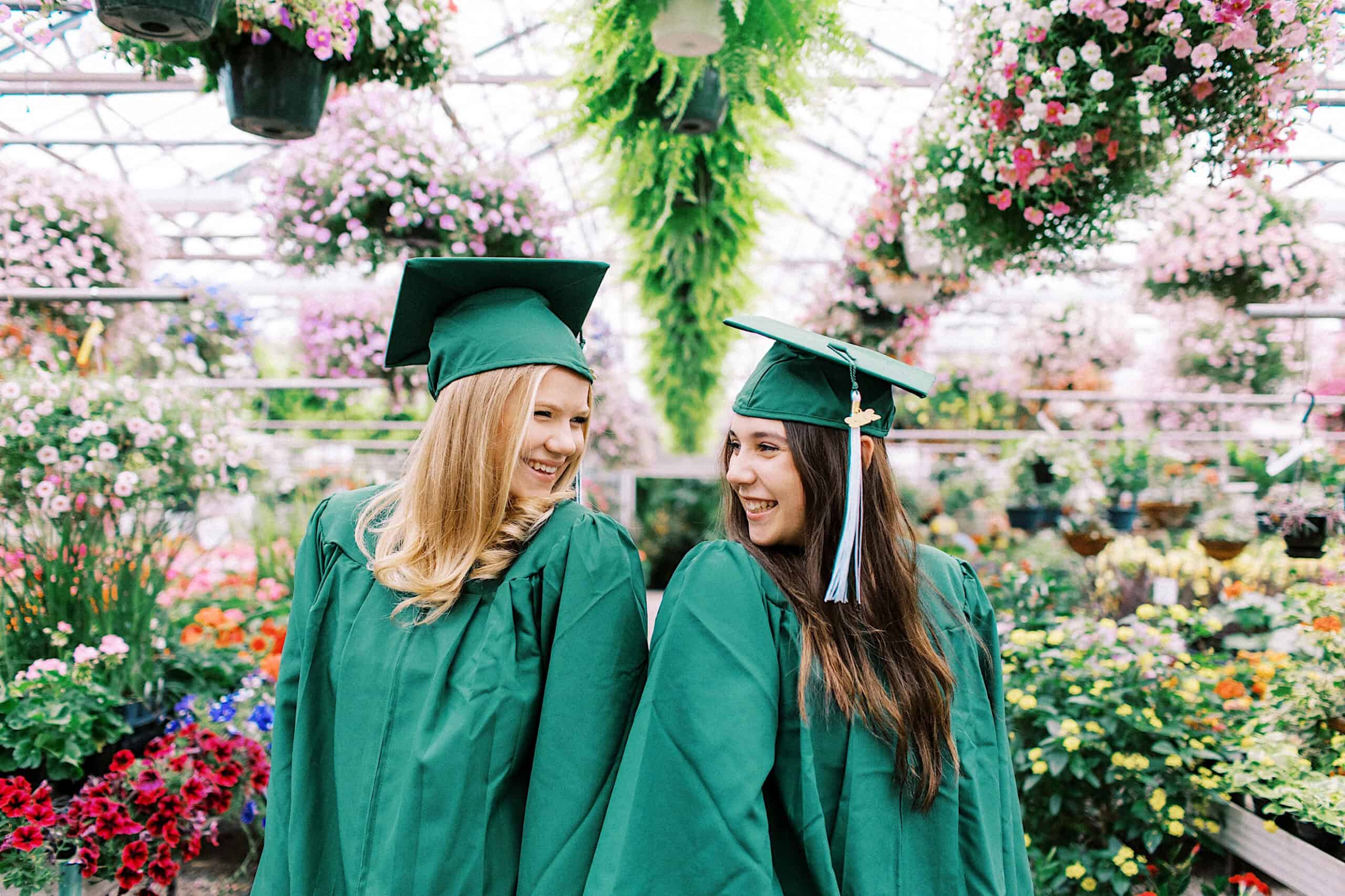 Two graduates in green caps and gowns stand together, smiling, surrounded by flowers and greenery in a greenhouse—capturing the joy of their Senior Portrait Session in Montgomery County.