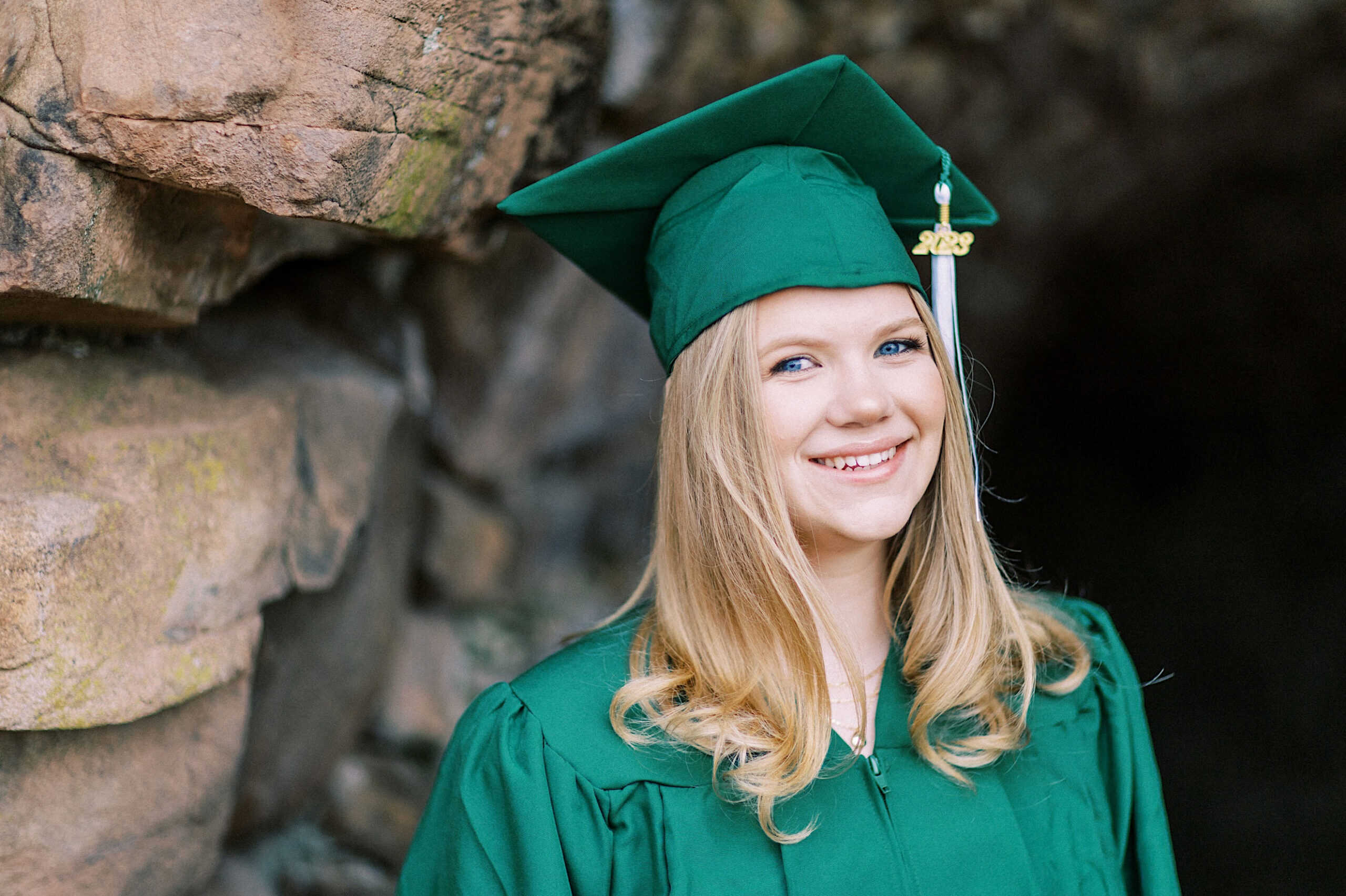 Young woman in a green graduation cap and gown standing in front of a rock wall, smiling at the camera during her Senior Portrait Session in Montgomery County.
