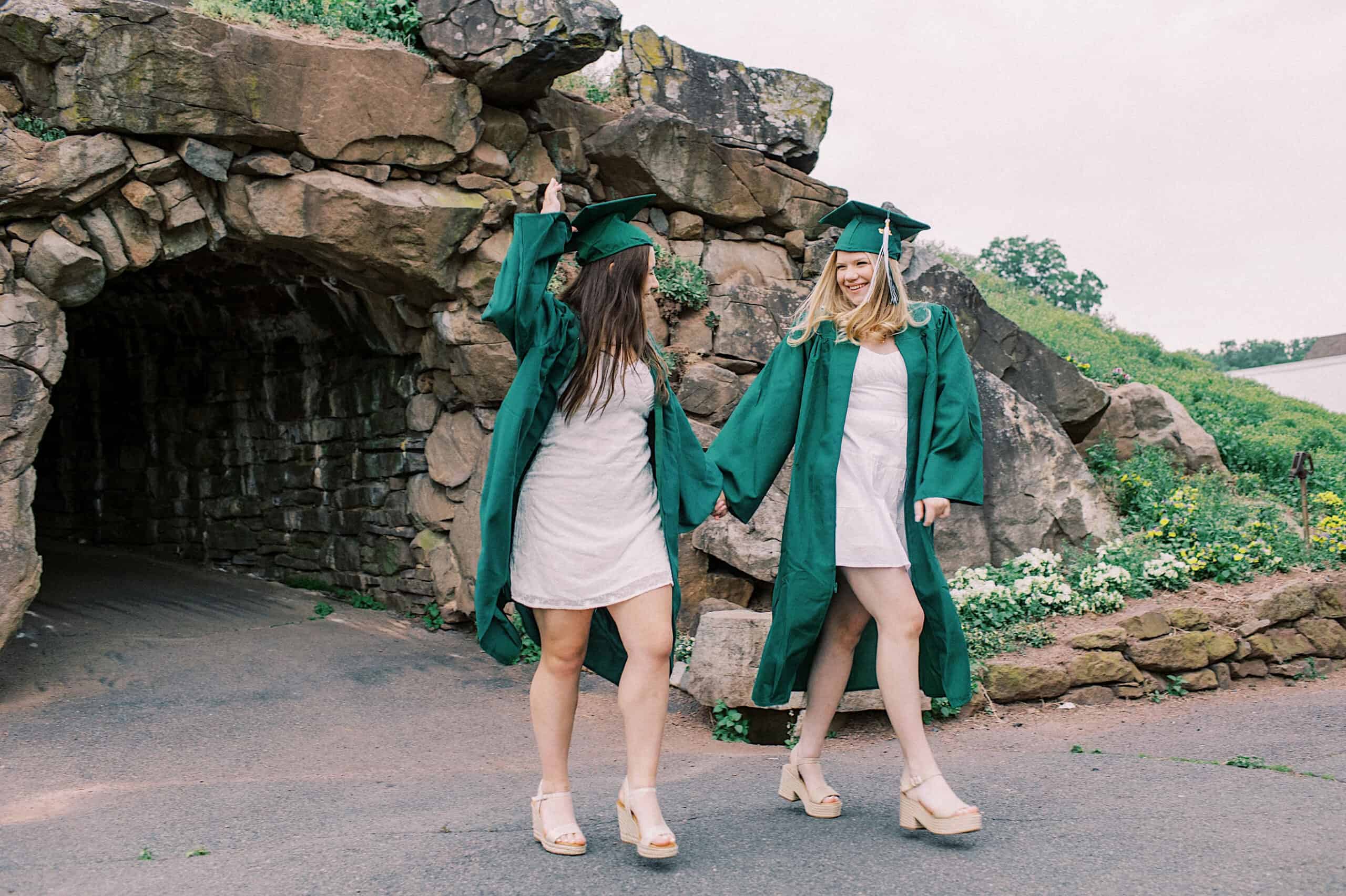 Two people in green graduation gowns and caps hold hands and smile while standing outside near a stone archway and greenery during their Senior Portrait Session in Montgomery County.