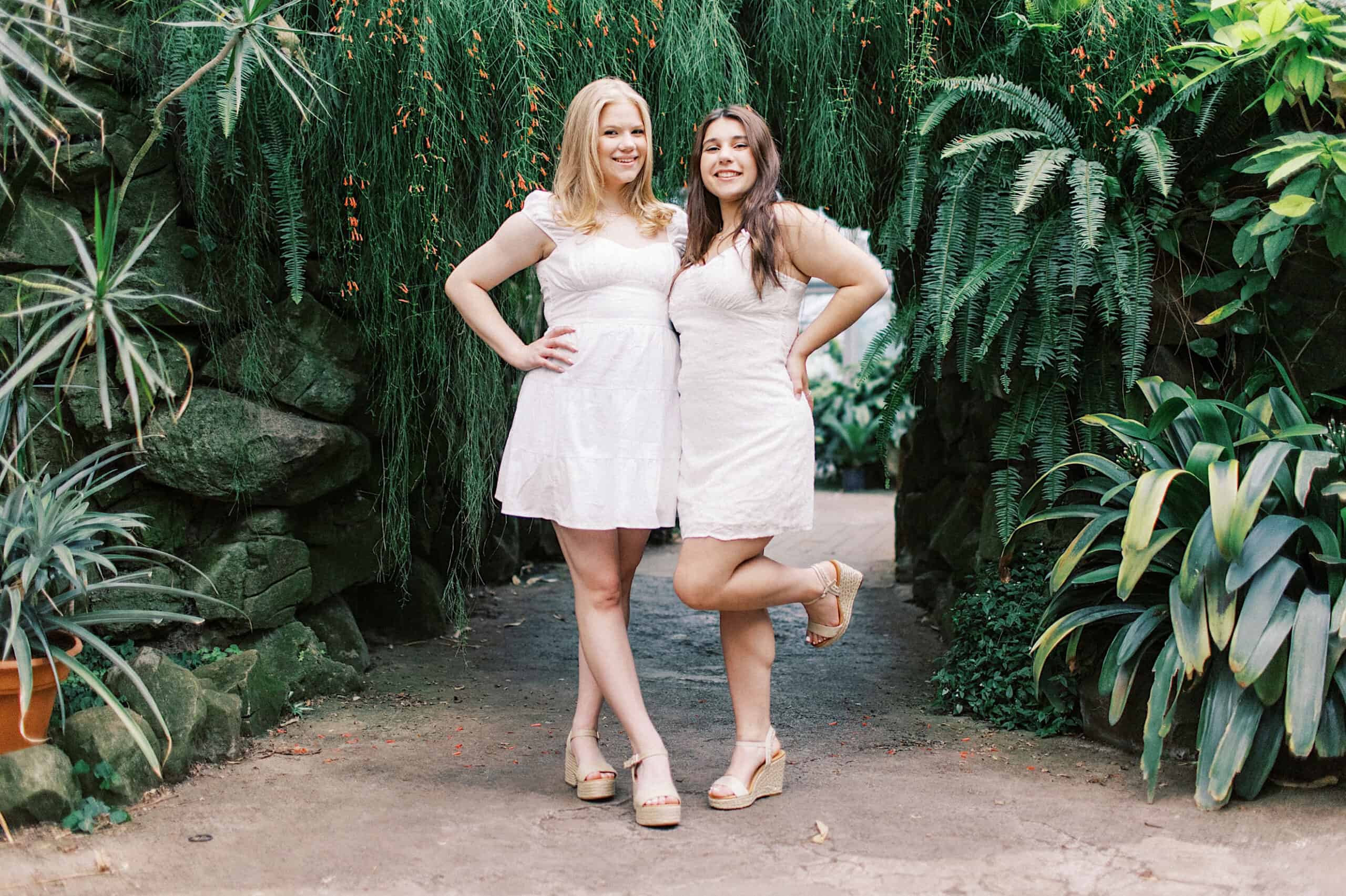 Two women in white dresses pose together outdoors during a Senior Portrait Session in Montgomery County, standing on a path surrounded by lush green plants and foliage.