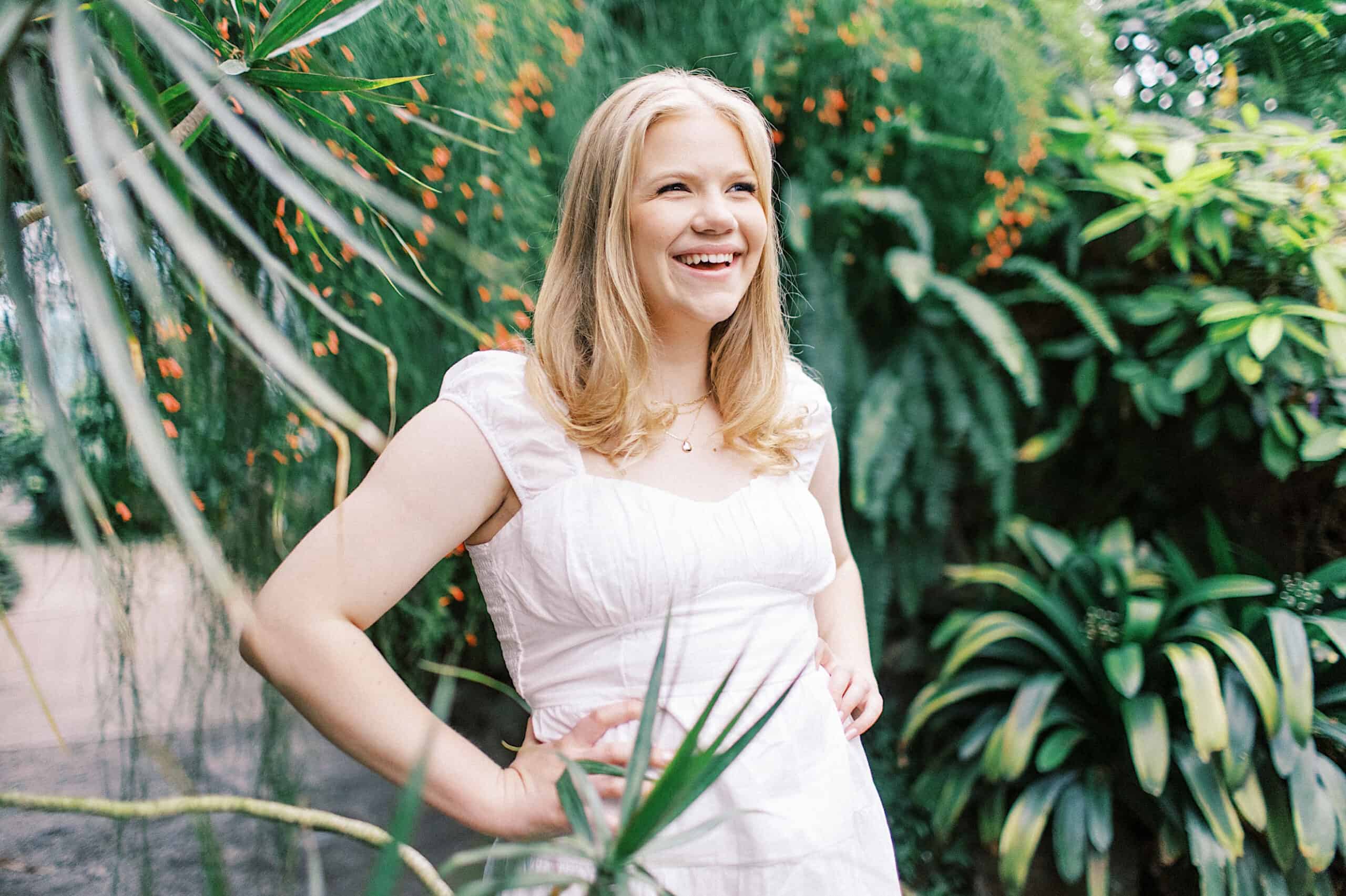 A woman in a white dress stands outdoors with hands on hips, smiling during her Senior Portrait Session in Montgomery County, surrounded by lush green plants and orange berries.