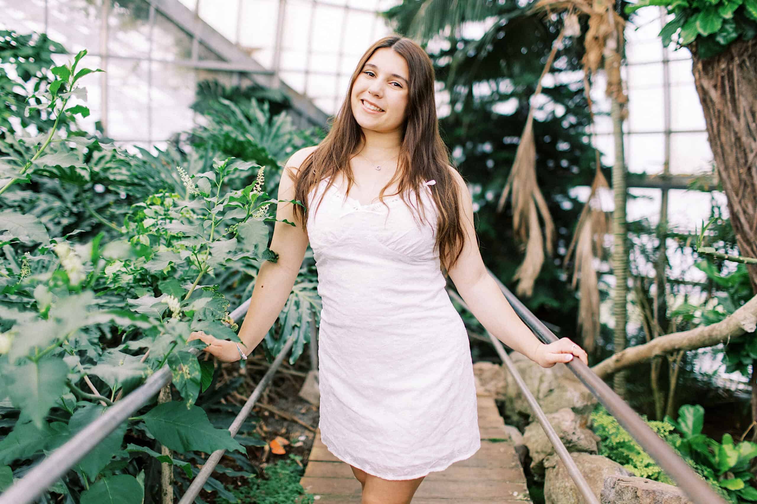 A young woman in a white dress stands on a path surrounded by green plants inside a greenhouse, smiling at the camera during her Senior Portrait Session in Montgomery County.