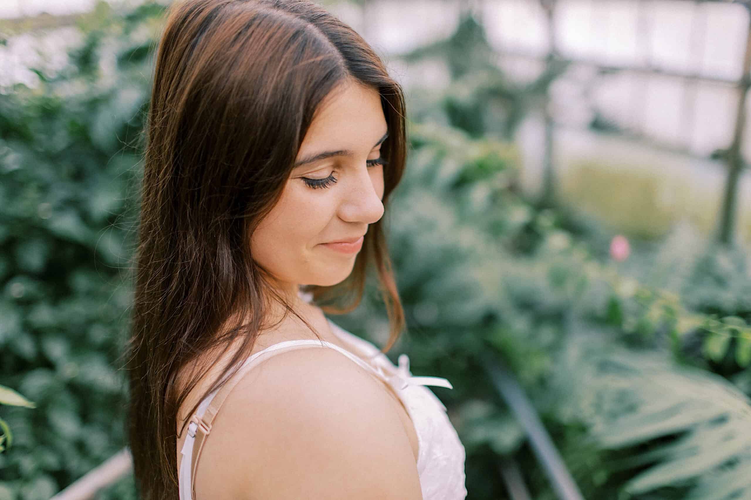 A young woman with long brown hair looks down and smiles softly while standing in a green, leafy environment during her Senior Portrait Session in Montgomery County.