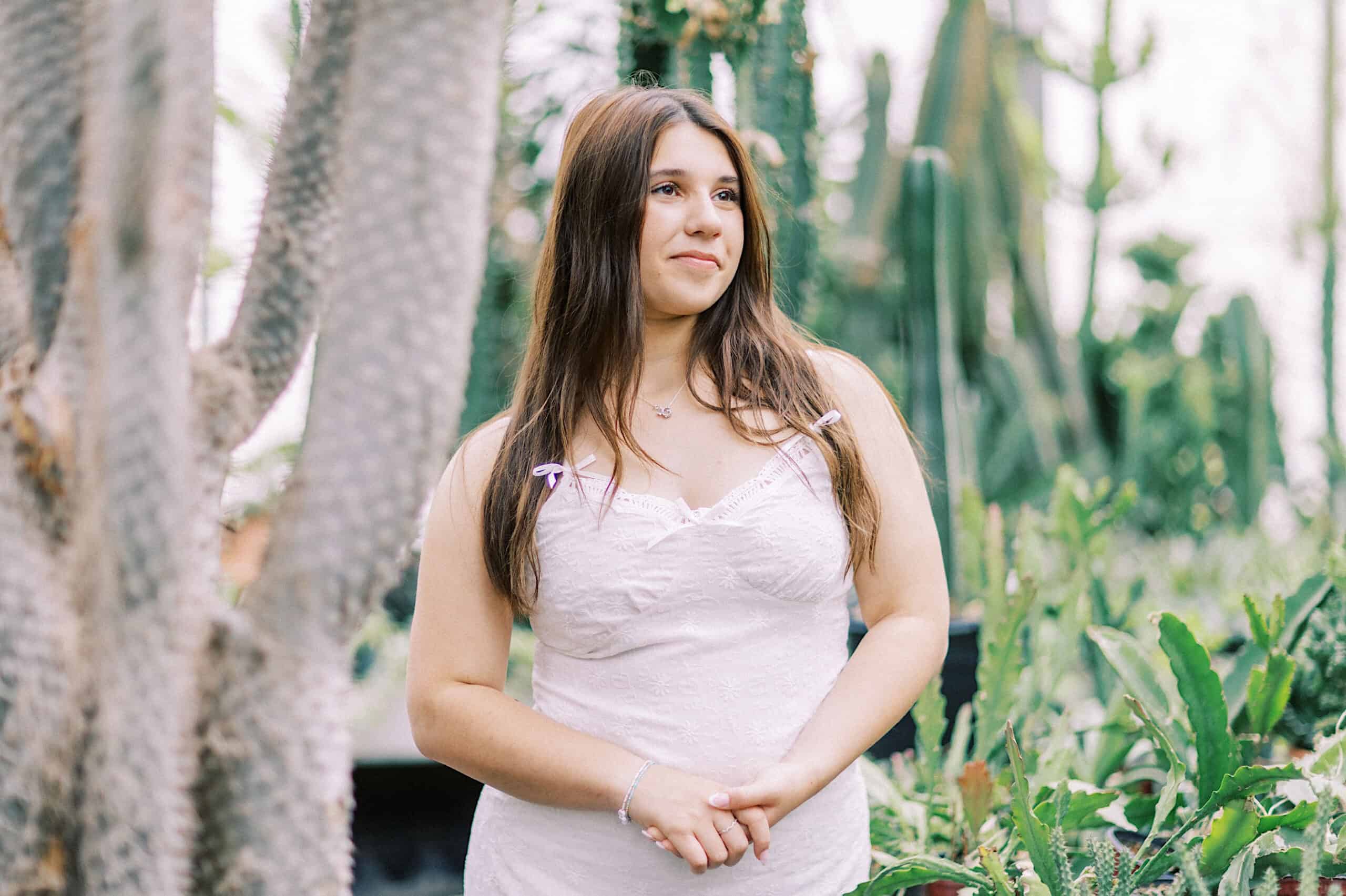 A woman in a white dress stands among green cacti and plants, looking slightly to the side with a neutral expression during a Senior Portrait Session in Montgomery County.